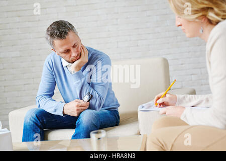 Sad male patient holding eyeglasses in hand and looking away desperately while his psychologist making necessary notes in medical card Stock Photo
