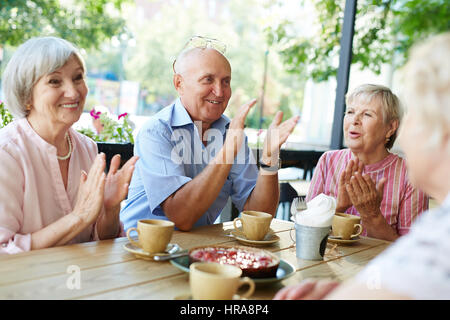 Group of smiling elderly people gathered together in outdoor cafe for tea-drinking and applauding joyfully Stock Photo