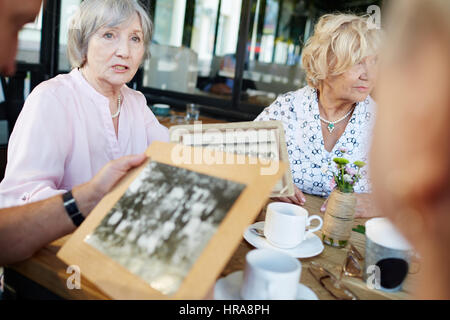 Elderly woman looking into distance thoughtfully while telling her friends story of her childhood and showing them black-and-white photos in outdoor c Stock Photo