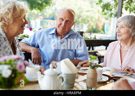 Senior friends having good time together in outdoor cafe, one woman turning over page of time-worn photo album Stock Photo