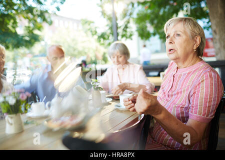 Portrait of elderly woman in striped summer dress drinking tea with her friends in outdoor cafe and looking into distance thoughtfully Stock Photo