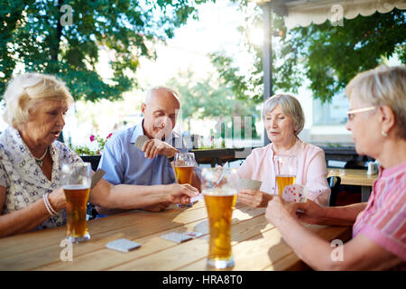 Group of four elderly people relaxing in cafe: they drinking beer and playing cards with interest Stock Photo
