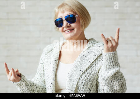 Trendy elderly woman in sunglasses making horn gesture with both hands and looking at camera with confident smile against white background Stock Photo