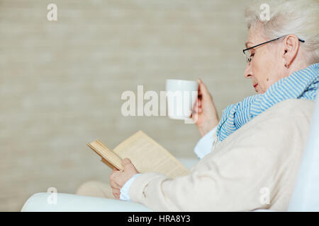 Grey-haired senior woman in blue-and-white scarf drinking cup of coffee and reading exciting book against beige background, profile view Stock Photo