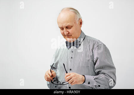 Waist-up portrait of bald senior man wearing striped shirt with bowtie looking sadly at eyeglasses in hands against white background Stock Photo