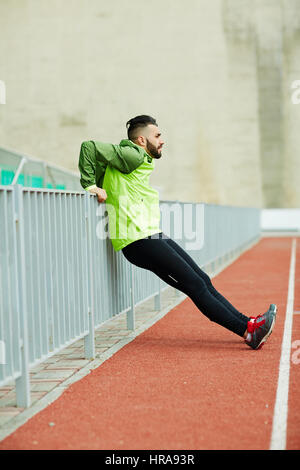 Side view image of young male sportsman warming up and stretching against rail before running practice in empty stadium Stock Photo