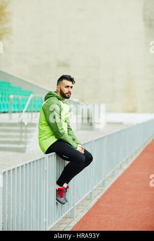 Young fashionable man sitting, resting on railings after running practice in stadium Stock Photo