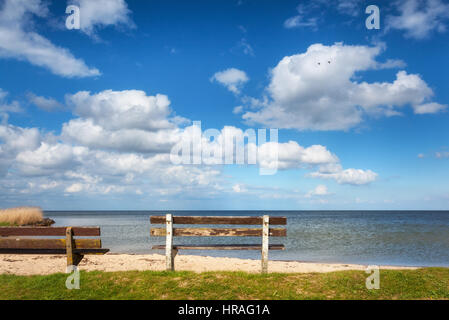 Bench on the sandy beach at the sea on the background of colorful blue sky with clouds at sunset. Beautiful landscape with empty old wooden bench Stock Photo