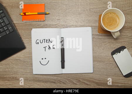 Open notebook with German text 'GUTEN TAG' (Good afternoon)  and a cup of coffee on wooden background. Top down view Stock Photo