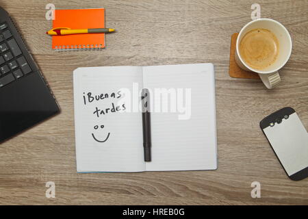 Open notebook with Spanish words 'Buenas Tardes' ( Good Afternoon) and a cup of coffee on wooden background. Stock Photo