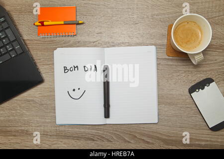 Open notebook with Portuguese words 'BOM DIA' (Good afternoon)  and a cup of coffee on wooden background. Top down view Stock Photo