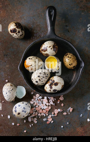 Whole and broken quail eggs with yolk in shell and pink sea salt crystal in small iron cast pan over old rusty texture metal background. Top view with Stock Photo