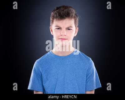 Teen boy making silly grimace - expressing disgust face. Upset child on black background. Emotional portrait of caucasian teenager looking at camera. Stock Photo