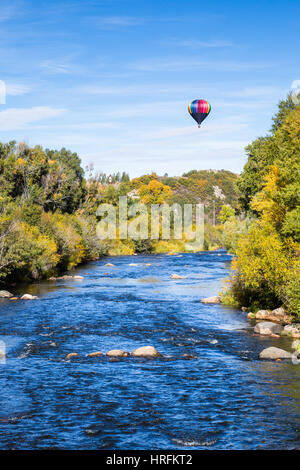 Trees are starting to turn yellow in autumn as a hot air balloon floats above a calm blue river near Steamboat Springs, Colorado. Stock Photo