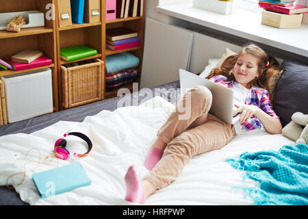 Pretty school girl lying on her bed, holding laptop and chatting with friends in social network Stock Photo