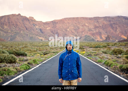 Portrait of sad man in blue jacket and hood standing on empty road and looking at camera on scenic mountains background Stock Photo