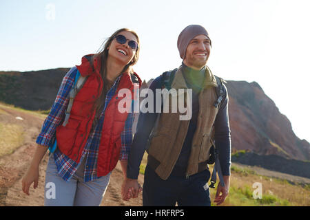 Portrait of smiling young couple wearing active clothes smiling brightly walking on hiking path in mountains together holding hands on sunny day Stock Photo