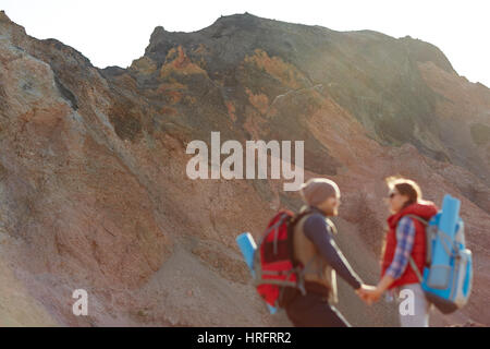 Couple of young travelers hiking in wild nature, man and woman wearing tourist gear with big backpacks standing holding hands against backgrounds of e Stock Photo