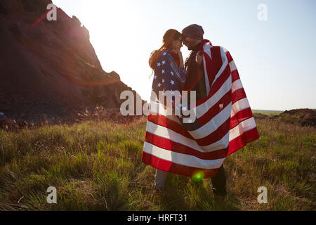 Romantic adventure of young couple standing on top of hill at sunset, wrapped in American banner and looking at each other enjoying mountain hike Stock Photo
