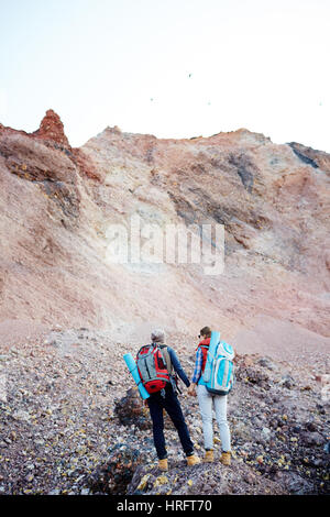 Rear view portrait of young couple, man and woman, wearing tourist gear and big backpacks standing on hill holding hands while traveling on hike in mo Stock Photo