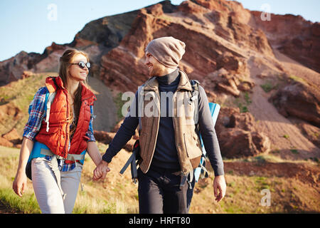 Portrait of young cheerful couple, man and woman, wearing tourist gear and big backpacks walking on hiking path through mountains holding hands, lit b Stock Photo