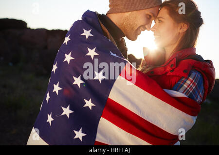 Side view portrait of tender romantic couple embracing wrapped together in big American flag at sunset Stock Photo