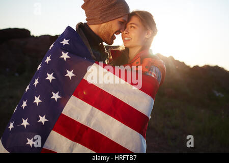 Portrait of young tourist couple standing close together in mountains, embracing tenderly wrapped in American flag at sunset Stock Photo