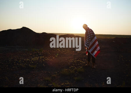 Backlit portrait of young fashionable man standing alone against sunset sky wrapped in big American banner taking a hike in mountains Stock Photo