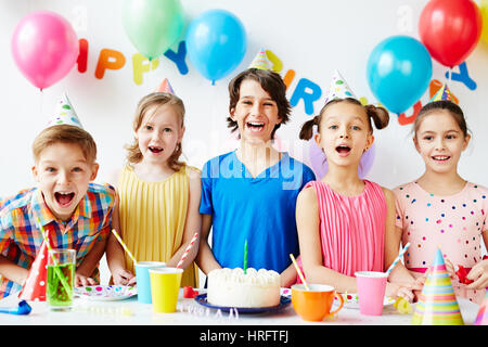 Portrait of five joyous children posing at birthday table with cake on it, smiling and showing their teeth with mouth open Stock Photo