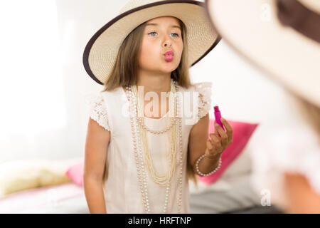 Mirror reflection shot of little girl playing dress up in mothers jewels and hat putting makeup and pink lipstick on, blowing air kisses and grimacing Stock Photo