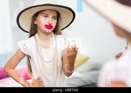 Mirror reflection shot of little girl playing dress up in mothers jewels and hat putting makeup and pink lipstick on, messing it up in smudges and loo Stock Photo