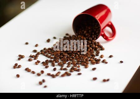 Background image:  Seeds of rich freshly roasted coffee spilling out of tipped red cup to white table Stock Photo