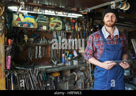 Portrait of handsome bearded mechanic wearing uniform overalls standing holding digital tablet against background of shelves filled with tools in work Stock Photo