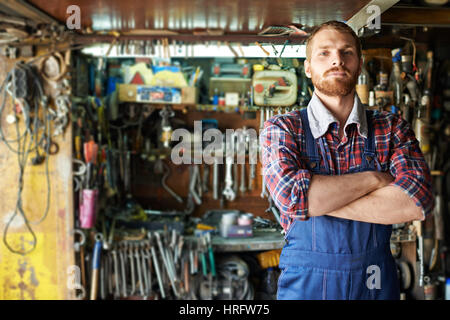 Portrait of handsome bearded mechanic standing confident with arms crossed  and looking at camera against shelves of tools and equipment in workshop Stock Photo