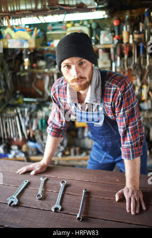 Portrait of handsome bearded mechanic wearing uniform overalls leaning on table   and looking confidently at camera against background of shelves fill Stock Photo