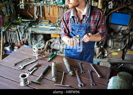 Unrecognizable bearded mechanic standing looking away and sorting working tools on table  in garage workshop Stock Photo