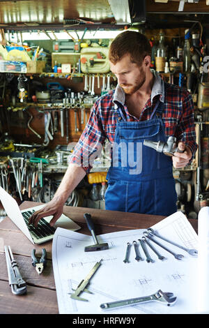 Portrait of handsome bearded mechanic wearing uniform overalls busy working in garage, assembling parts using tools and blueprints while looking down  Stock Photo