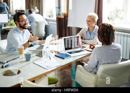 Multi-ethnic group of two businesspeople and one businesswoman sitting at table in meeting room and discussing details of their new project, modern of Stock Photo