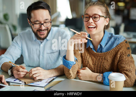 Waist-up portrait of young female employee with wide smile having project discussion while sitting next to her male colleague in eyeglasses Stock Photo