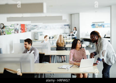 Interior of busy open plan office: male and female workers sitting at their desks, Afro-American superior discussing business project with pretty Asia Stock Photo