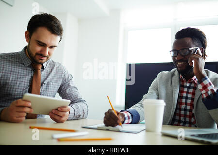 Bearded middle-aged businessman creating presentation on digital tablet while his Afro-American colleague distracted from work due to personal call Stock Photo
