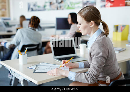 Busy open plan office: pretty middle-aged interior designer sitting at desk and making sketch with help of pencil, her colleagues wrapped up in work Stock Photo