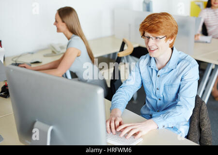 Young red-haired employee looking at computer screen with smile while writing response email to his business partner Stock Photo
