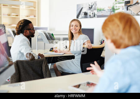 Portrait of beautiful blond-haired worker looking at camera with toothy smile while sharing ideas concerning business project with her male colleagues Stock Photo