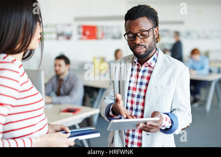 Concentrated Afro-American businessman in eyeglasses holding digital tablet in hand and pointing at something on its screen, his female colleague list Stock Photo