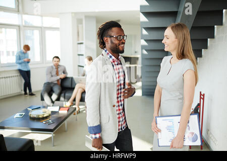 Having break after productive business meeting: Afro-American employee and his pretty female colleague chatting on foreground while other workers seen Stock Photo