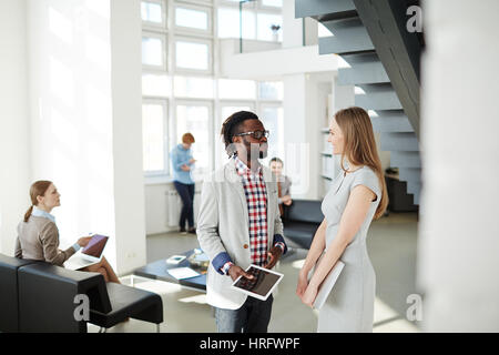 Pretty businesswoman with laptop standing next to Afro-American colleague with digital tablet while having informal meeting in office lobby Stock Photo