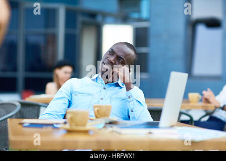Portrait of pensive African American businessman looking away in daydream while relaxing at outdoor table with laptop on it during lunch break at work Stock Photo
