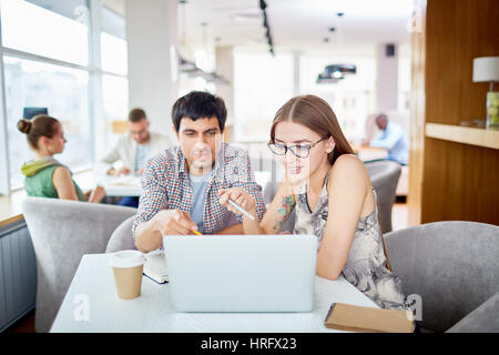 Portrait of two young people, man and woman,  working with laptop at table in modern cafe, pointing at screen and discussing ideas Stock Photo