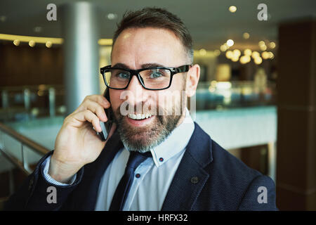 Portrait of cheerful mature businessman smiling while making phone call from modern airport lounge Stock Photo